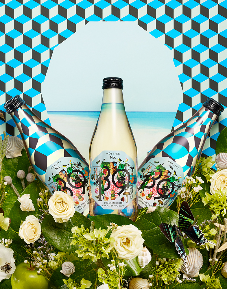 THREE WHITE CIDER BOTTLES ON GEOMETRIC PATTERNED BACKGROUND SURROUNDED BY SUMMER THEMED FLOWERS SHOT BY DAVID FILIBERTI